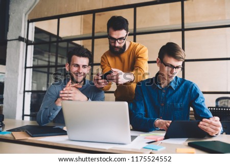 Millennial hipster guys collaboration together in coworking space sitting at desktop with laptop while one of them making photos of computer screen, man reading sms message via digital tablet