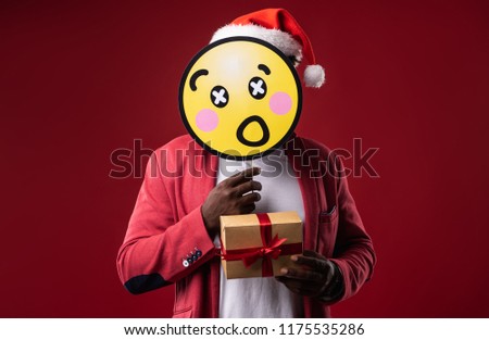 I am hiding. Man in santa hat covering face with emoji and demonstrating gift box with ribbon. Isolated on red background