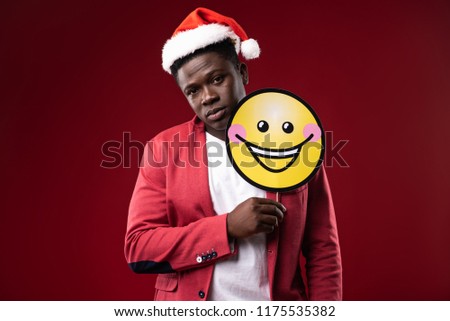 Cheer up. Handsome man in santa hat posing with emoji icon. Isolated on red background