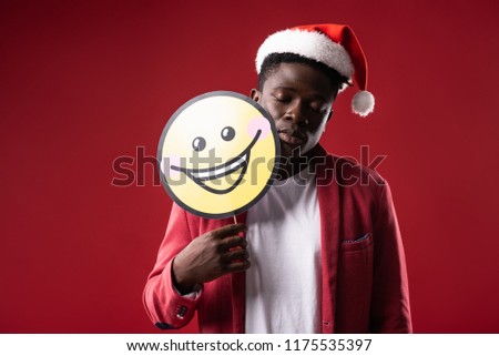 Need some sleep. Handsome man in santa hat posing with carton icon of smiling face. Isolated on red background