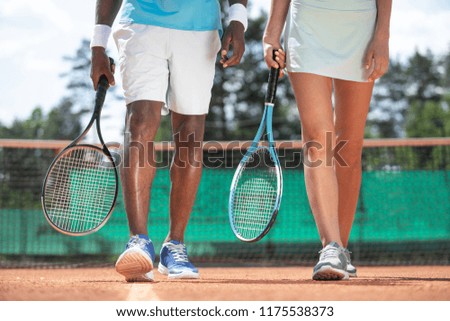 Close up of male and female legs while playing tennis in open air. Couple is walking along playground during break. They are holding rackets