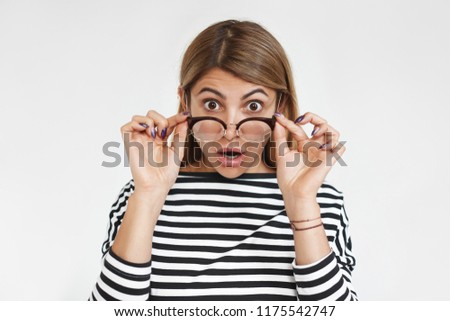 Picture of amazed funny young woman in stylish white top with black stripes staring at camera with mouth opened and eyes popped out, lowering her trendy eyeglasses, shocked with big sale prices