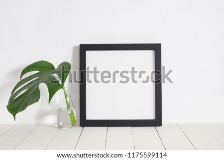 Black square frame with green monstera leaf in vase on wooden table