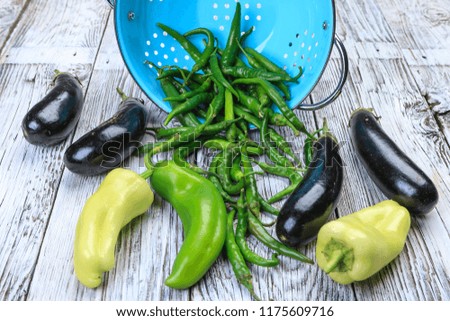 An overview photo of eggplant and various peppers in a blue colander and on a wooden board.