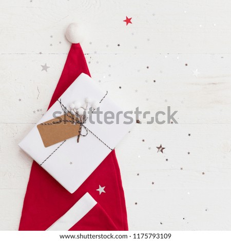 Closeup of Santa's hat and beautiful Christmas gift wrapped in white paper and decorated with tag on white wooden table. DIY present idea. Holidays concept.