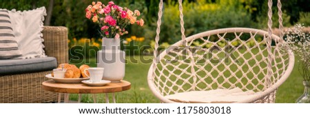 Flowers on table between hanging chair and rattan settee in the garden. Real photo