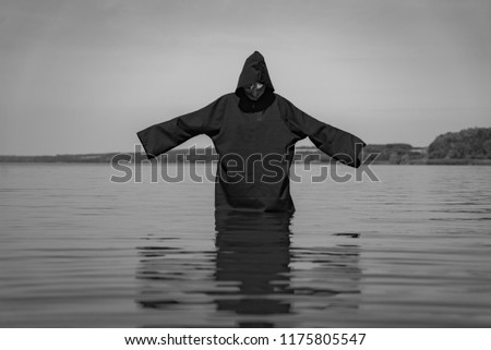 a ghost in a black cloak stands in the lake with hands raised on the background of nature. The phantom is depicted in a black and white photo