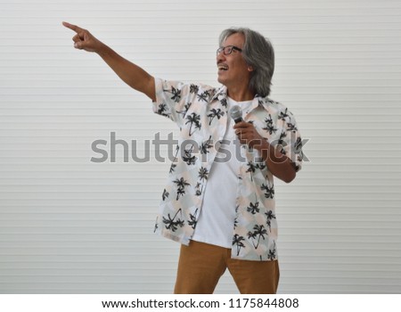 Happy senior traveler asian man wearing glasses, summer shirt and brown shorts speaking with microphone and pointing finger up over white wall background, Business summer holiday concept