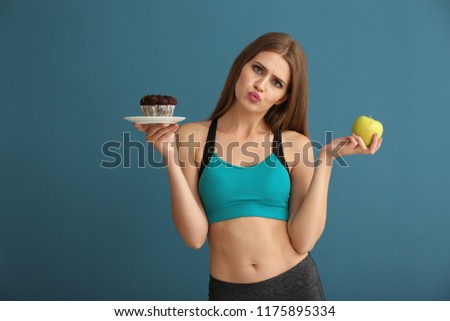 Young woman in sportswear choosing between cake and apple on color background