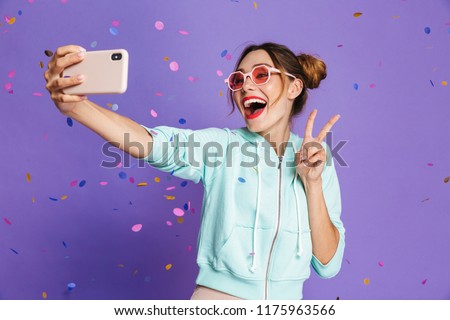Portrait of a happy young girl with bright makeup isolated over violet background, taking a selfie under confetti shower