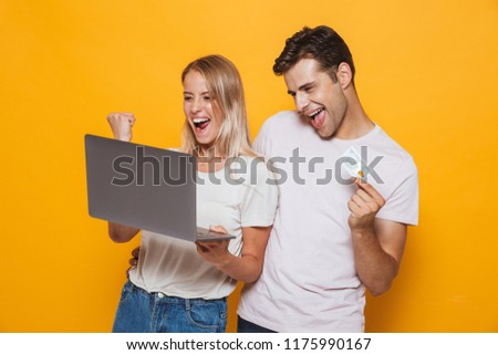 Photo of happy excited young loving couple isolated over yellow wall background using laptop computer make winner gesture holding credit card.