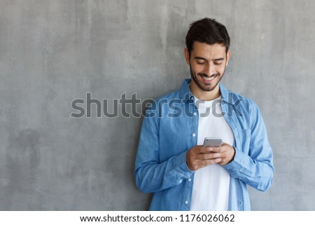 Indoor picture of European Caucasian man isolated on gray background dressed in blue denim shirt and typing messages on his smartphone communicating with friends in social networks, smiling happily