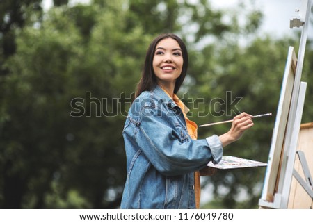 Street portrait of a young brunette asian woman standing and painting in the city park outdoor, with blurred bushes and trees in the background. Education, nature, hobby, lifestyle. Labor of love
