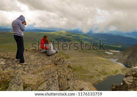 A photographer makes a photo of a loving couple is standing on the Altai mountains kissing against the background of a valley bathed in sunlight, lakes with glaciers, rocks with snow and white clouds.