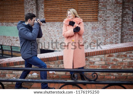 Man takes picture of woman in peach-colour warm coat against red bricks wall outdoor.