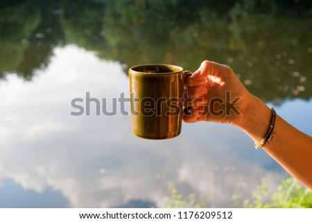 Female hand holding a cup in a green wooded and lake landscape background