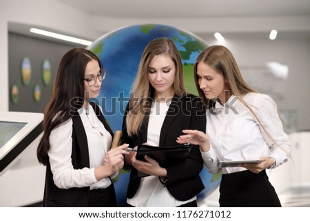 Three beautiful young smiling girls in business clothes. A team of professional colleagues. White shirts, black suits and glasses. Modern office with globe. Ecology and environmental protection.
