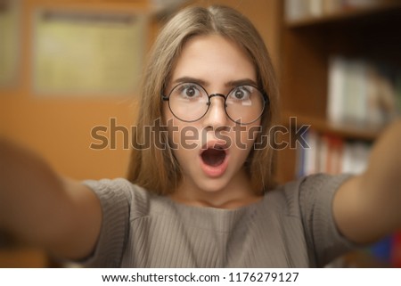 Portrait of a shocked young student schoolgirl lady with long hair wearing eyeglasses standing in empty classroom looking camera take a selfie.