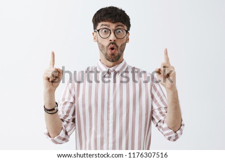 Excited enthusiastic good-looking mature guy with beard folding lips and staring with excitement and amazement at camera while pointing up with raised hands finding something interesting