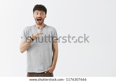 Excted and surptised pleased dark-haired guy with beard cannot control happiness standing with super star smiling with dropped jaw, looking and pointing right with amazement and joy Royalty-Free Stock Photo #1176307666
