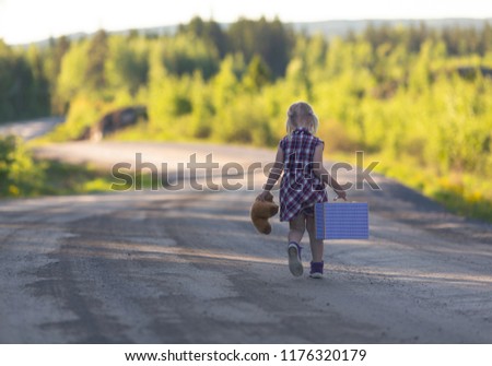 Caucasian girl walking away alone on an empty highway with bear and suitcase. Concept image of a runaway child with loneliness and sad feelings. Image has a vintage effect. Royalty-Free Stock Photo #1176320179