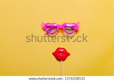 Holiday minimalistic flat lay: funny pink sunglasses and masquerade plastic lips on stick. Yellow background.