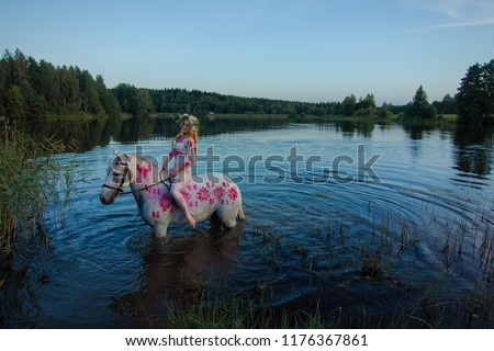 Blonde girl in white and red bodypain bathes with a horse in the lake at sunset.
