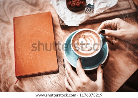 Cup of coffee latte and red book on wooden table or background in woman hands from above.