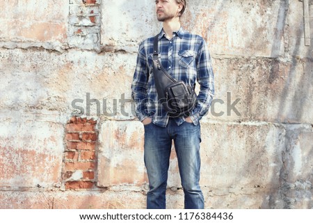 Young hipster man in casual wear with stylish leather crossbody bag standing against concrete wall. Modern city lifestyle.  Royalty-Free Stock Photo #1176384436