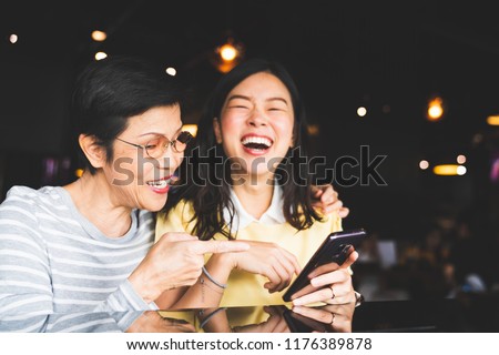 Asian mother and daughter laughing and smiling on a selfie or photo album, using smartphone together at restaurant or cafe, with copy space. Family love, holiday activity, or modern lifestyle concept Royalty-Free Stock Photo #1176389878