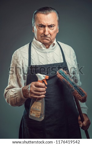 Senior old man disgusted by the idea of cleaning, holding cleaning chemicals and wearing apron Royalty-Free Stock Photo #1176419542