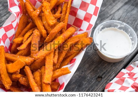 Paper basket of sweet potato fries with salt and a side of ranch dressing on rustic wooden table