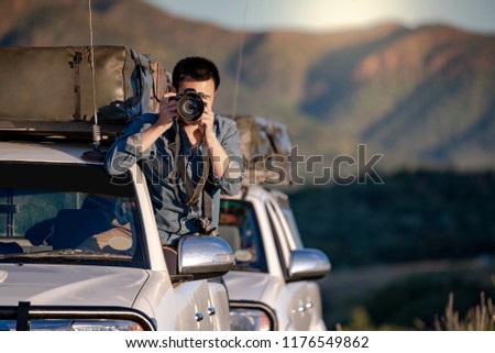 Young Asian male traveler and photographer sitting on the car window taking photo on road trip in Namibia, Africa. Travel photography concept