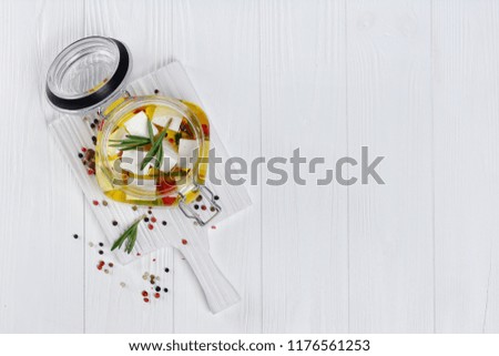 Marinated feta cheese with olive oil and spice of red chili pepper and rosemary in glass jar on white wooden board with copy space