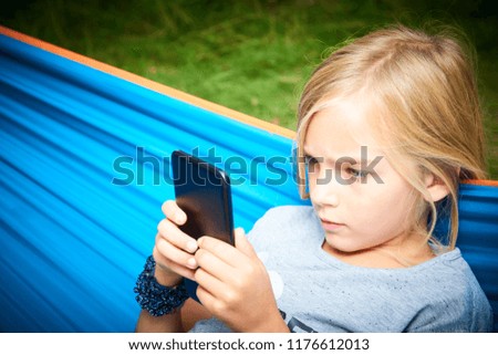 Child blond girl playing with a smart phone while lying on hammock outdoors 
