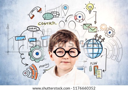 Cute little caucasian boy in glasses wearing shirt looking at camera standing near concrete wall with colorful management and business leadership icons. Future of business concept. Toned image