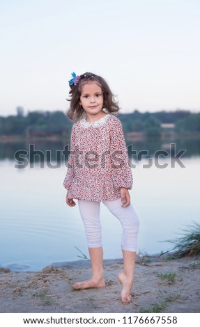 The fashionable girl in white trousers near the lake. The child costs barefoot on sand. The beautiful little girl poses near a reservoir