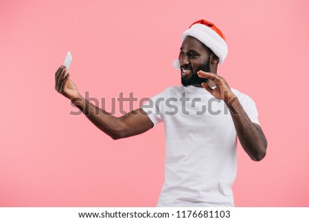 smiling african american man in santa claus hat with smartphone waving isolated on pink 