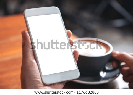 Mockup image of hands holding white mobile phone with blank screen while drinking coffee in modern cafe