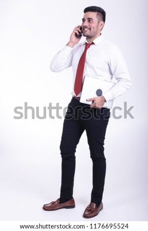 Businessman isolated. Handsome young indian businessman holding laptop, using a phone talking, confident looks. Full length shot.