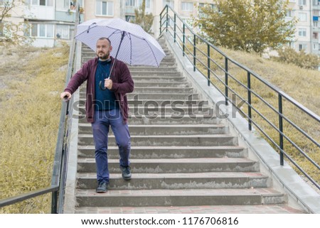 An unshaven man of 35-40 years old in a sweatsuit descends the stairs under an umbrella.