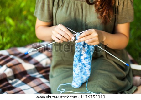 A young woman sitting in a city park on green grass and knitting a blue sweater with knitting needles on a summer day. Close Up. Get Creative everywhere