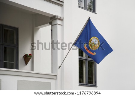 Idaho flag. Idaho state flag hanging on a pole in front of the house. State flag waving on a home displaying on a pole on a front door of a building. Flag raised at a full staff.