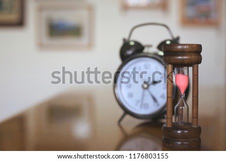 A close-up shot of a brown hourglass on a living room table. Alarm clock is the background selective focus and shallow depth of field