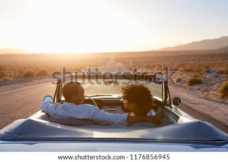 Rear View Of Couple On Road Trip Driving Classic Convertible Car Towards Sunset Royalty-Free Stock Photo #1176856945