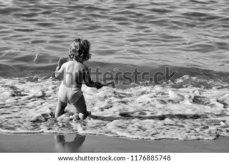 Small curious blonde child boy standing on sea coast beach with wavy water sunny twilight outdoor playing on natural background, horizontal picture