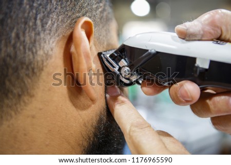Barber trim African man client with clipper machine in barbershop.Professional trimmer tool cuts beard and hair on young black guy in barber shop salon. Male beauty treatment process in close up
