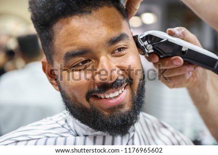 Barber shop pictures. Happy young black man sitting in a barber's chair. Professional barber shaving a client Royalty-Free Stock Photo #1176965602