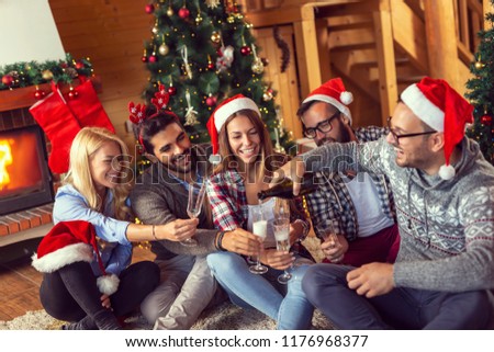 Group of friends sitting on the floor next to a nicely decorated Christmas tree, pouring champagne into glasses and making a toast
