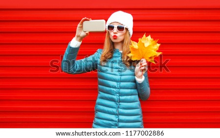 Fashion autumn pretty woman takes a picture self portrait on a smartphone with maple leaves on red background